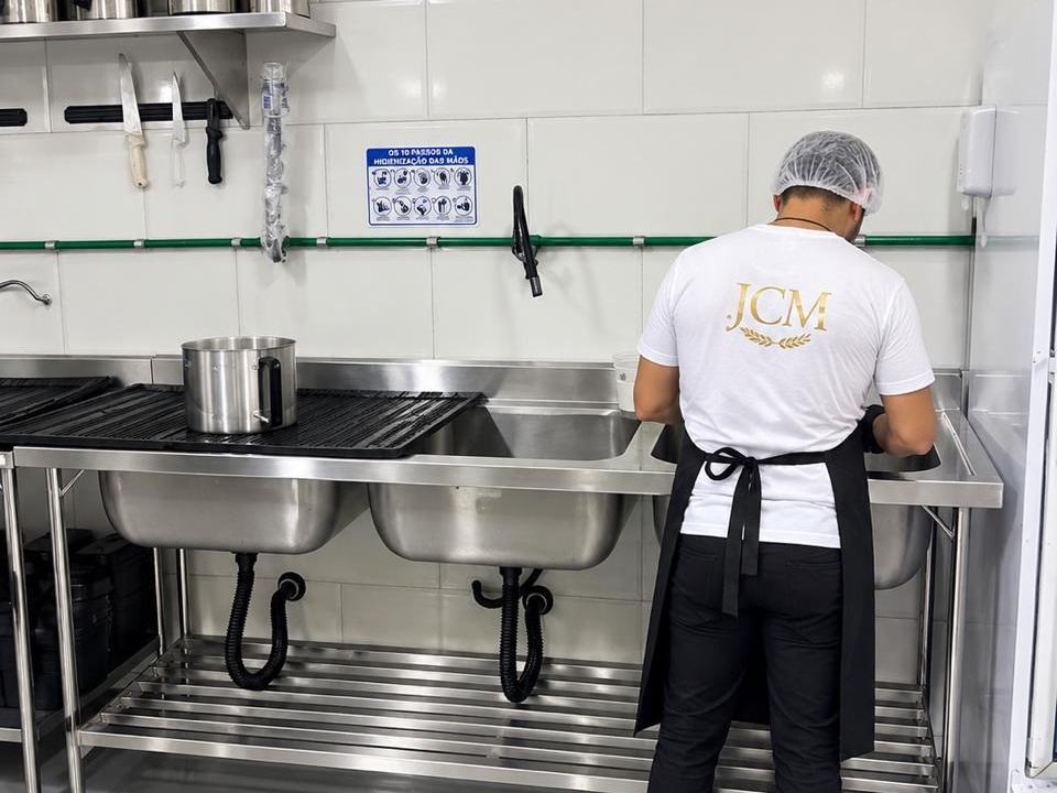 Funcionário da JCM de touca higienizando utensílios na pia